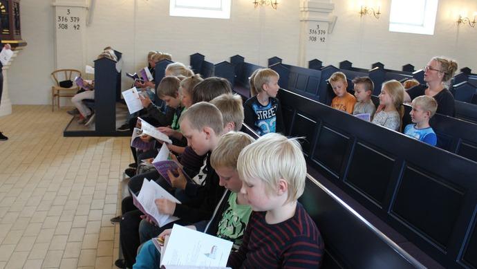 Børn sidder i en kirke og holder bøger, mens de lytter opmærksomt til en prædiken.
