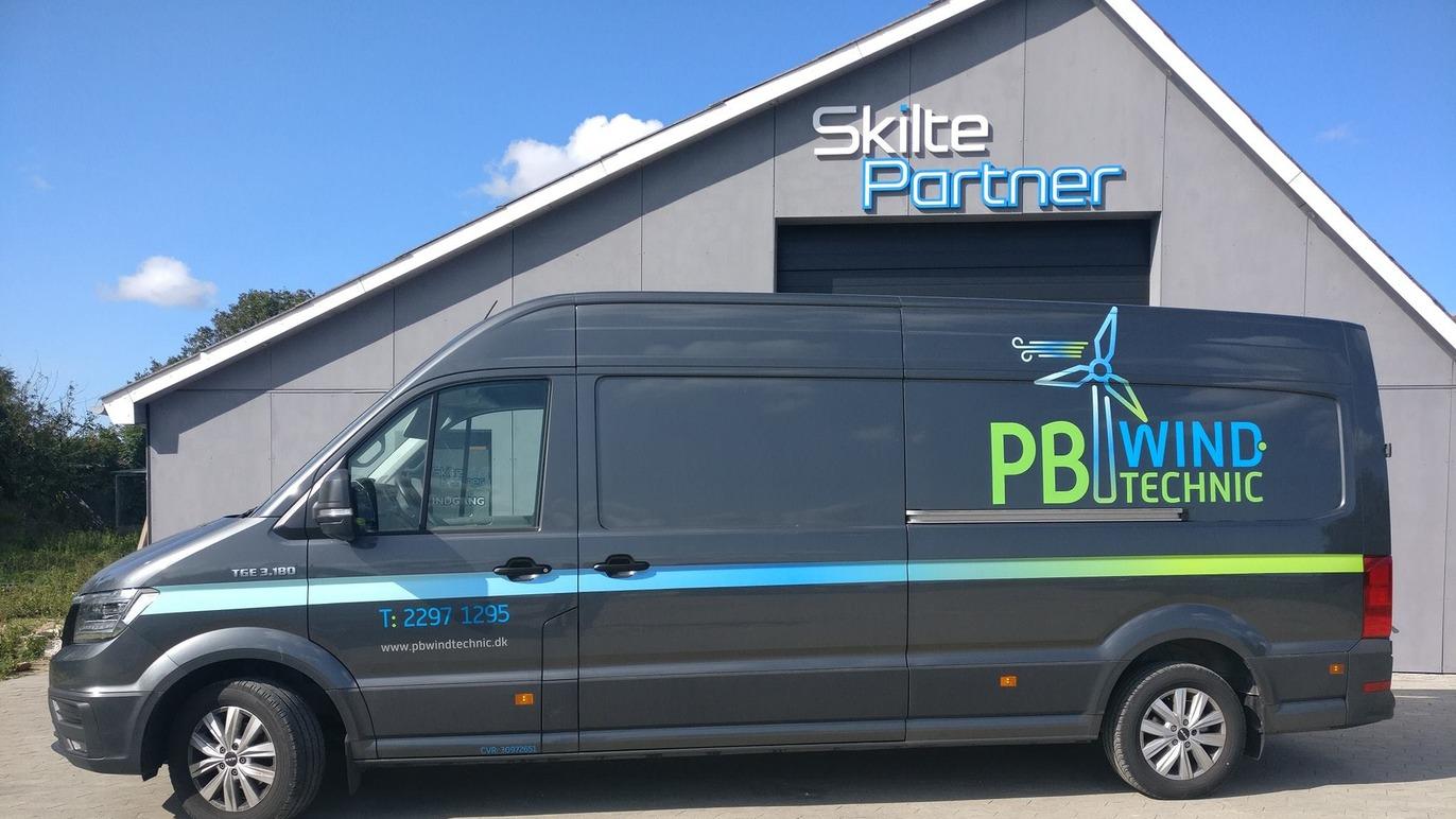 En grå varevogn med teksten "PB Wind Technic" parkeret foran en bygning med "Skilte Partner" på facaden.