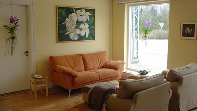 Stue med orange sofa, maleri af blomster på væggen og store vinduer, der giver naturligt lys.