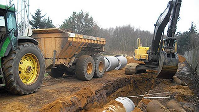 En gravemaskine arbejder på en byggeplads med rør og en dumper, der står ved siden af.