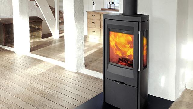 Moderne brændeovn med flammer, placeret på et lyst trægulv i et stilrent rum.