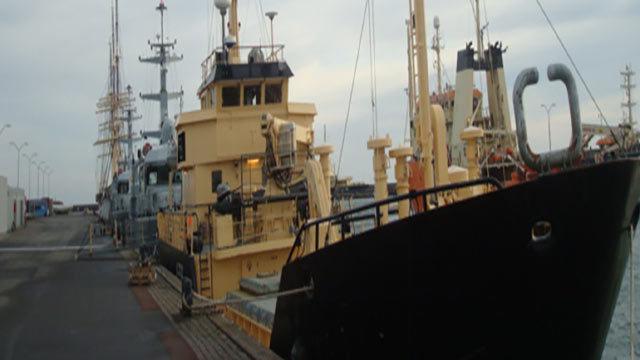 En stor fiskerbåd ligger ved kajen med flere andre skibe i baggrunden under en grå himmel.