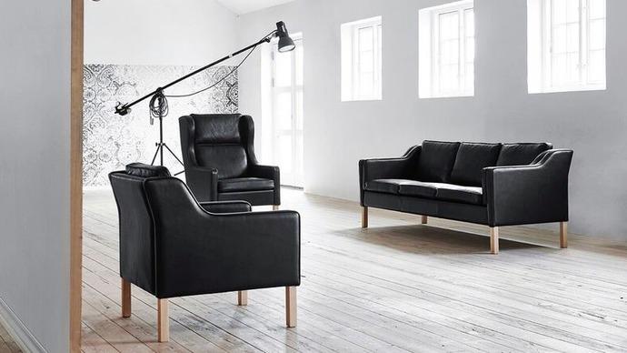 Moderne stue med sort lædersofa og lænestol, træben og store vinduer, der giver naturligt lys.