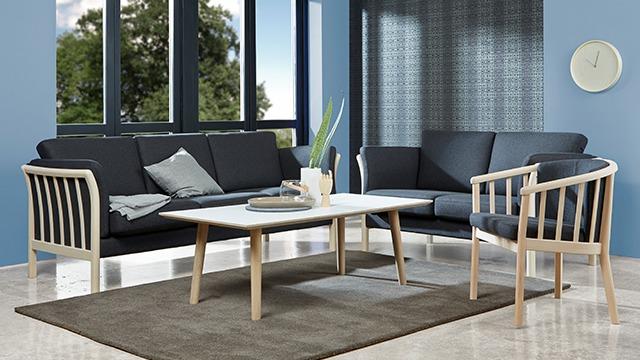 Moderne stue med en sofa, lænestol og et bord i lyst træ, omgivet af store vinduer og en blød tæppe.