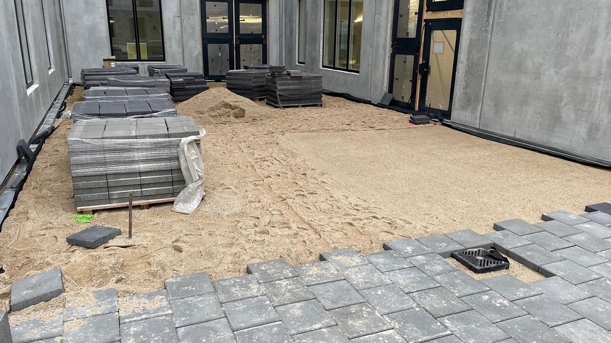 Billede af en byggeplads med grå fliser og sand, der ligger klar til at blive lagt på en gård.