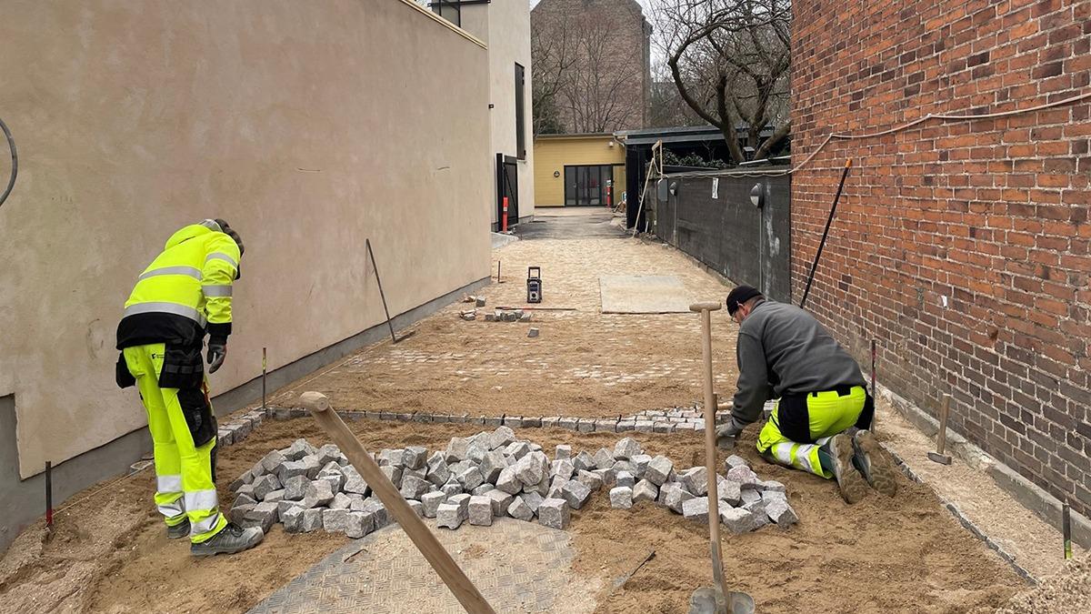 To arbejdere lægger sten på en ny belægning i en smal gang mellem bygninger.