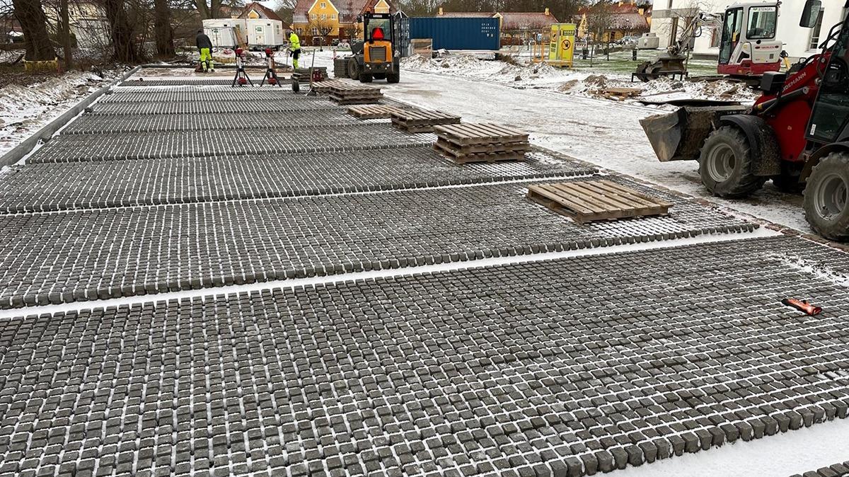 Byggeplads med metalrør og paletter på jorden, maskiner og arbejdere i baggrunden.