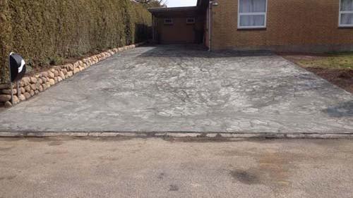 Ny betonindkørsel foran et hus med en garage og en hæk langs kanten.