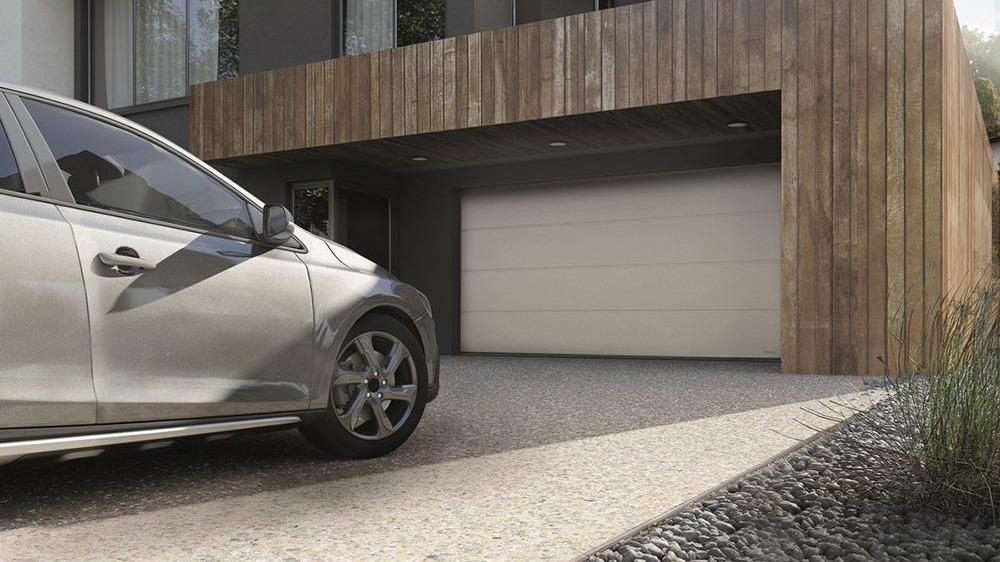 En sølvfarvet bil parkeret foran en moderne garage med træfacade og en hvid garageport.