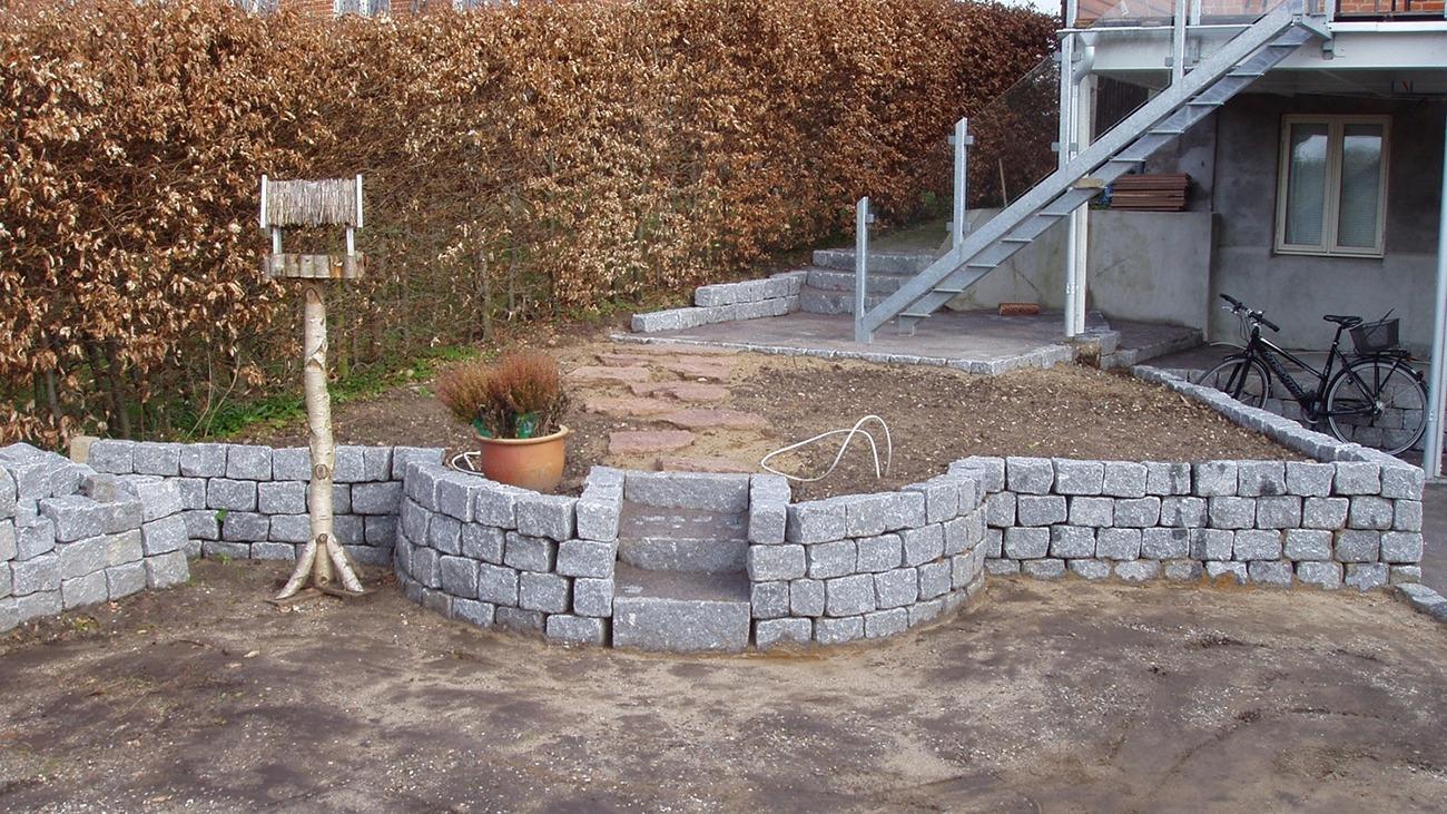 En have med en stenmur, en fuglefoder, og en trappe, omgivet af buske og en cykel ved siden af huset.