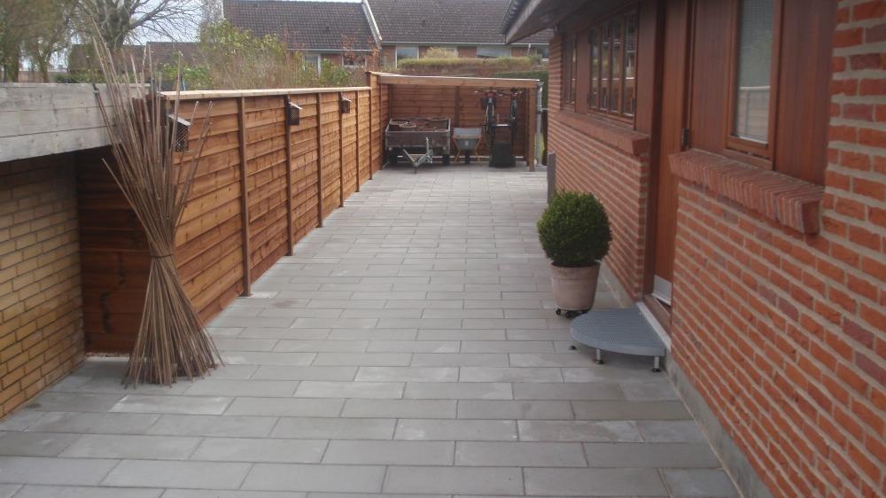 En flisebelagt terrasse med en træhegn og en plante i en krukke ved siden af en murstensvæg.