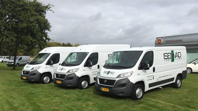 Tre hvide varevogne parkeret på en græsplæne med træer i baggrunden. Varevognene har logoer på siden.