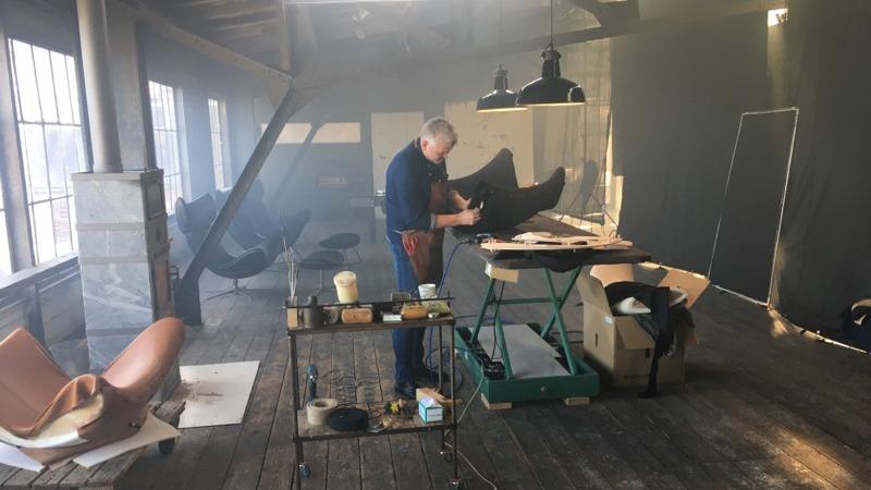 En håndværker arbejder med læder i et lyst, røgfyldt værksted med moderne møbler.