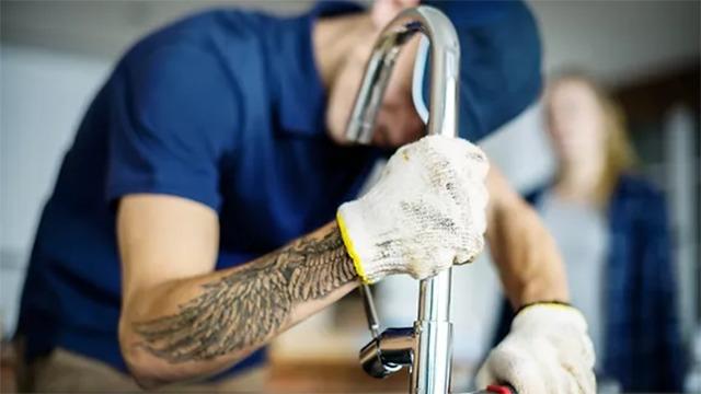 En håndværker installerer en vandhane med handsker på, mens en person observerer i baggrunden.