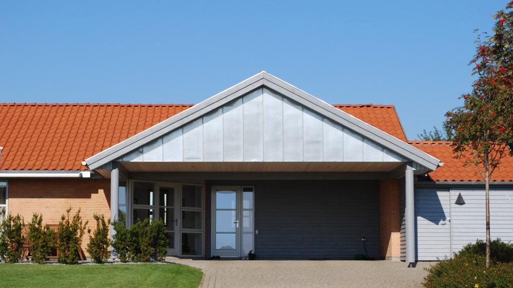 Moderne hus med rød tag og grå facade, indgang med glasdør og grøn græsplæne foran.