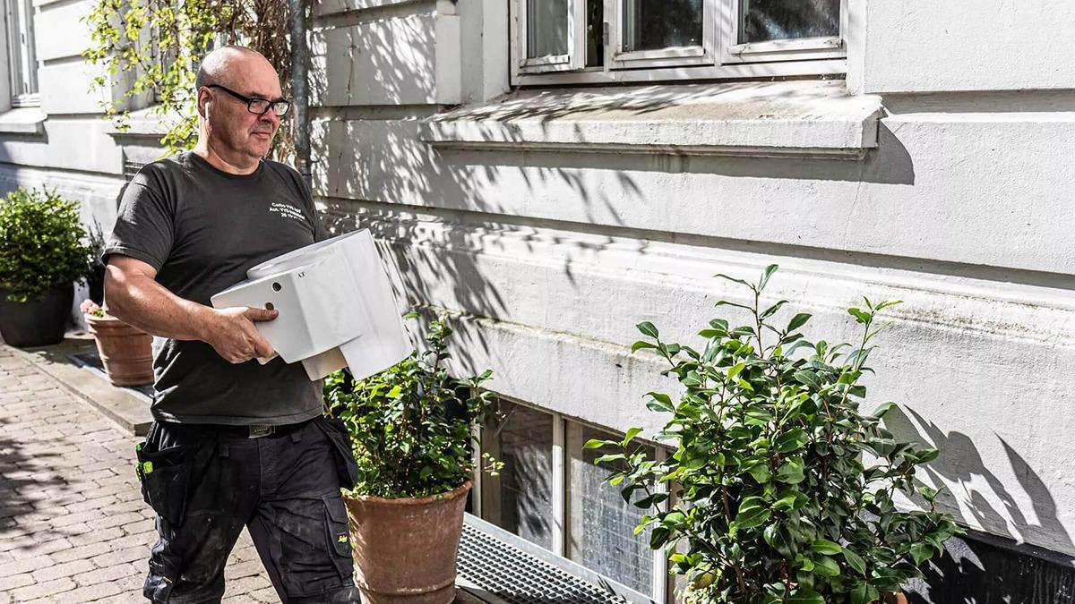 En mand bærer en kasse og går forbi en bygning med planter i potter.