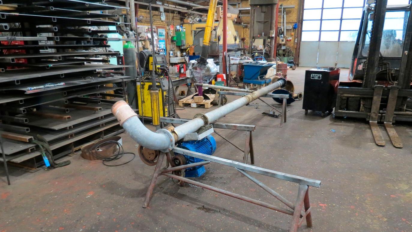 Et langt rør med en blå pumpe står i en værksted med metalplader og værktøj i baggrunden.