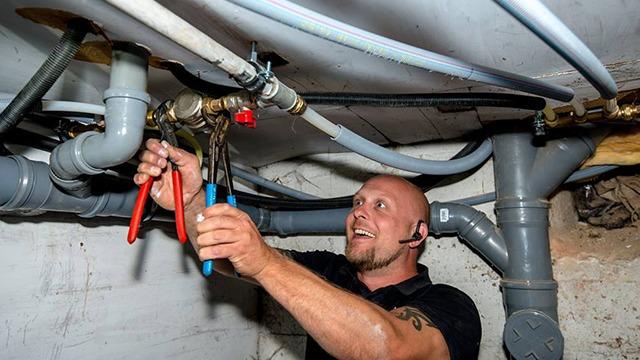 En håndværker arbejder med rør og værktøj under en vask i en kælder.