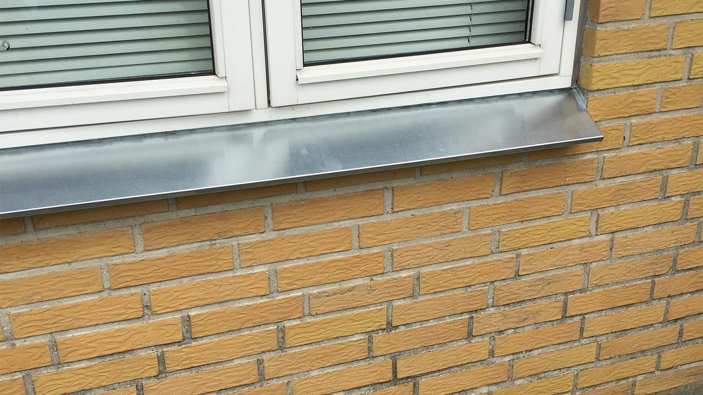 Sølvfarvet vindueskarm i metal på en murstensvæg med gule mursten.