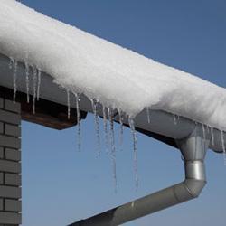 Tagrende med smeltende sne og istapper under en sneklædt tagkant mod en blå himmel.