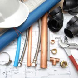 Værktøj og rør til VVS-arbejde, herunder kobberrør, fittings og en skruenøgle på en byggeplan.