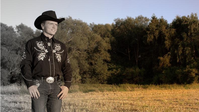 En cowboy i sort skjorte med broderi står på en mark med træer i baggrunden under en klar himmel.