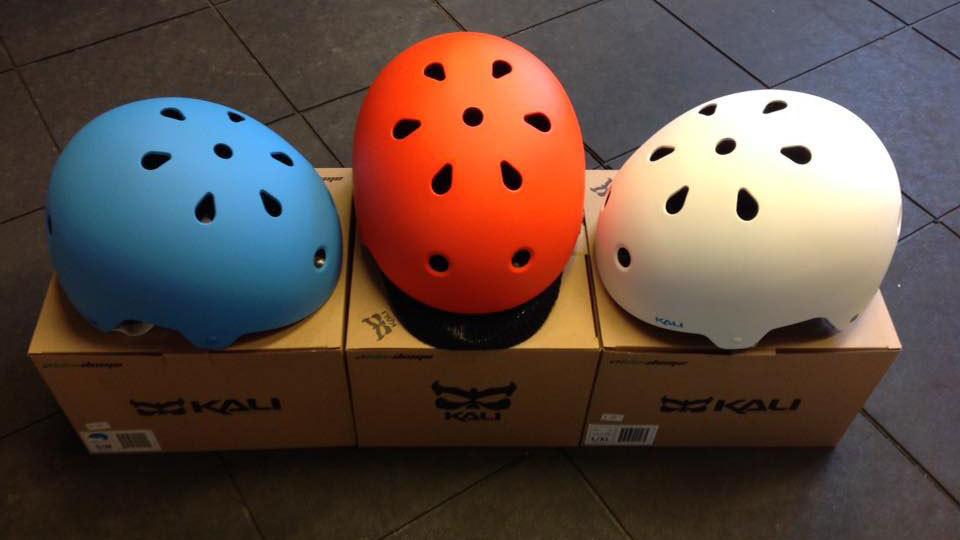 Tre cykelhjelme i blå, rød og hvid farve, placeret på papkasser i en butik.