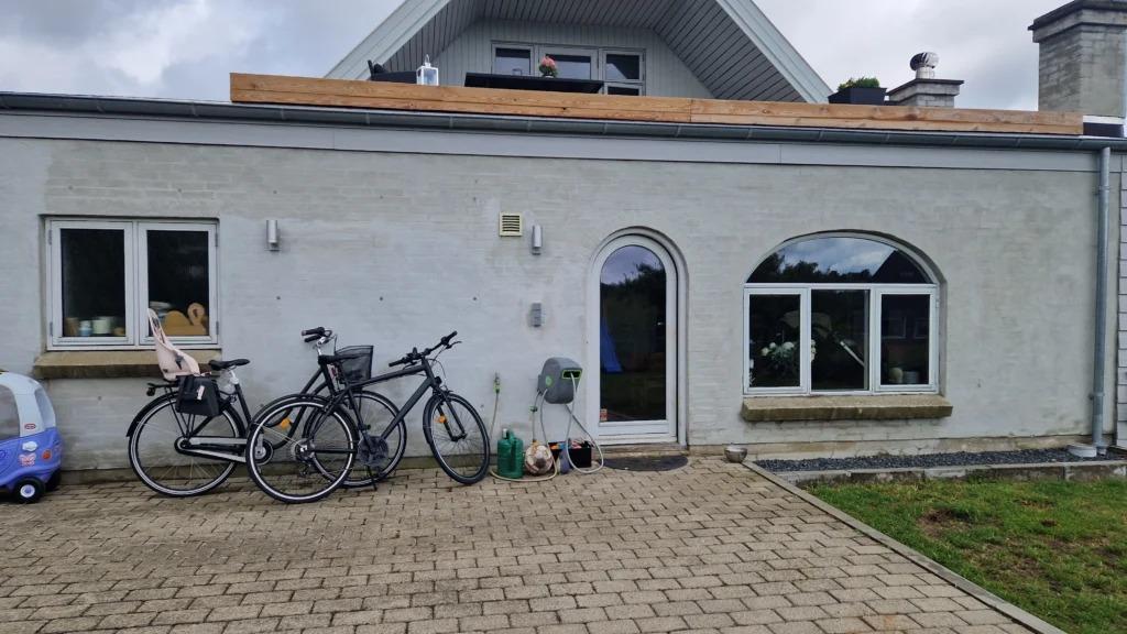 En grå murstenshus med to cykler parkeret foran og en indgangsdør, omgivet af en stenbelægning.