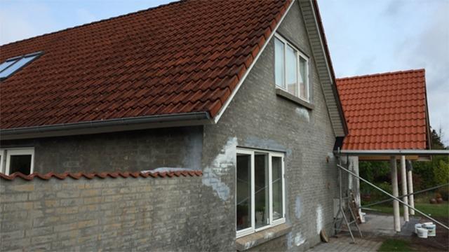 Hus med grå vægge og rød tagsten, der er under renovering, med stillads og malerudstyr synligt.