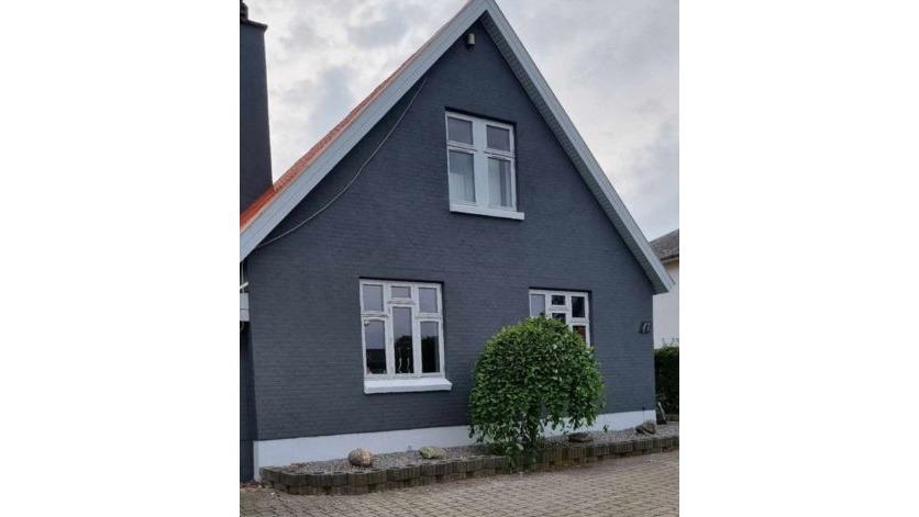 Et gråt hus med hvidt vindue og en busk foran, beliggende på en stille gade.
