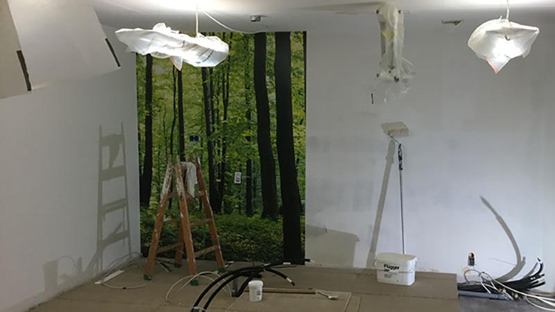 Indendørs rum med malerarbejde, en stige, og en vægdekoration af en skov. Kabel og malingsudstyr er synligt.