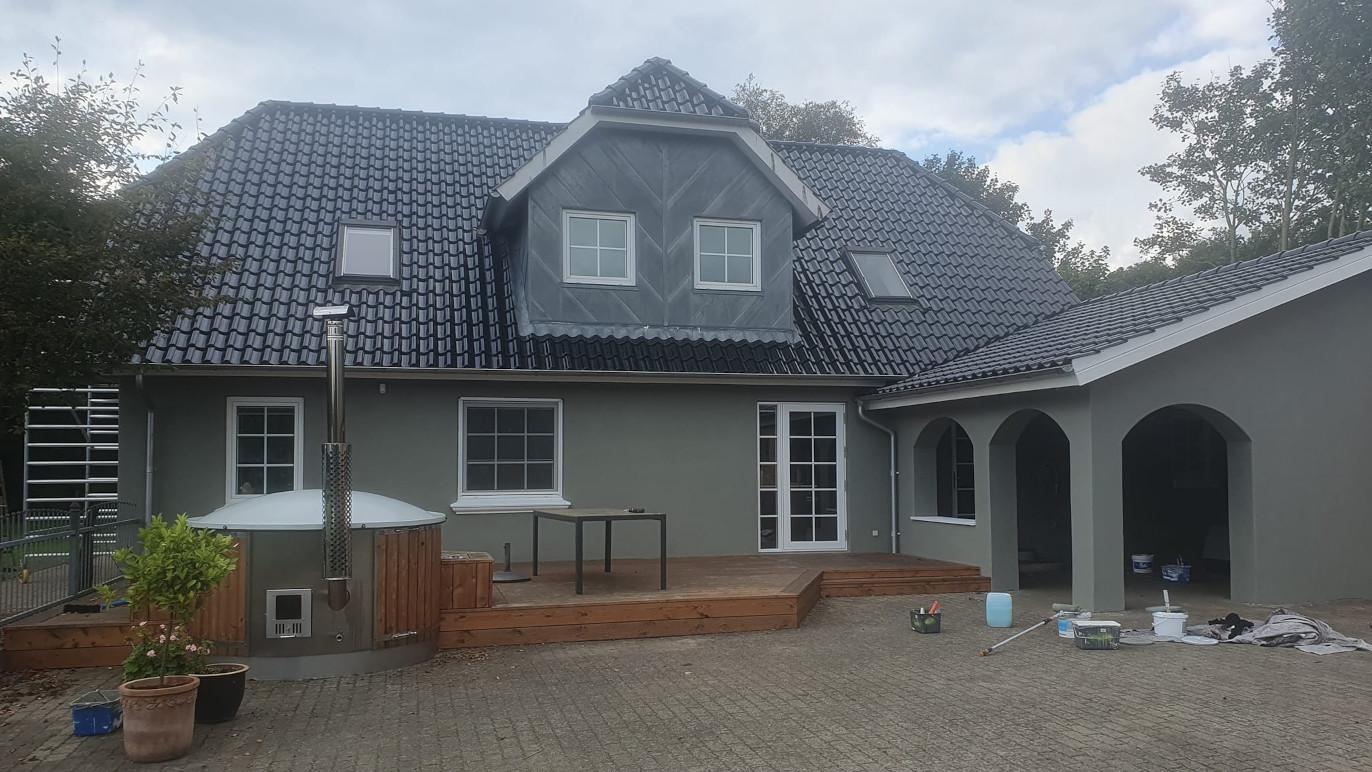 Et moderne hus med grå vægge, sort tag og en terrasse, omgivet af træer og en gårdsplads.