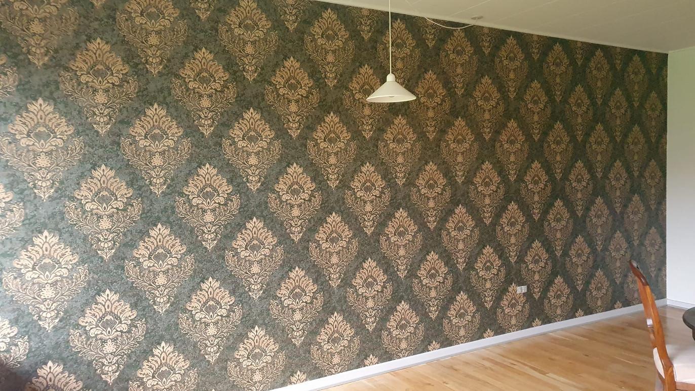 Væg med mønstret tapet i brune og gyldne nuancer, belyst af en hvid lampe, med trægulv og en stol i forgrunden.
