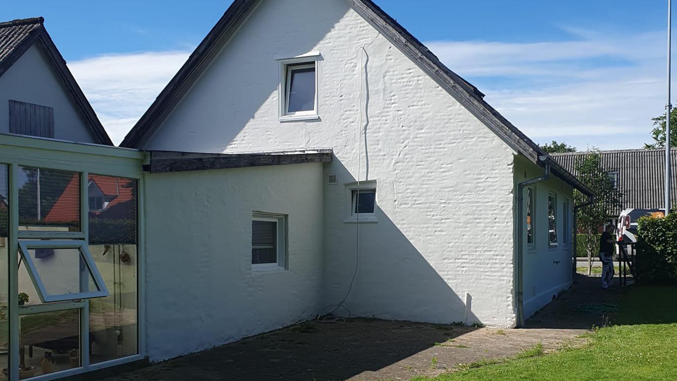 Hus med hvid facade og skråtag, beliggende i et roligt område med blå himmel og grønne planter.