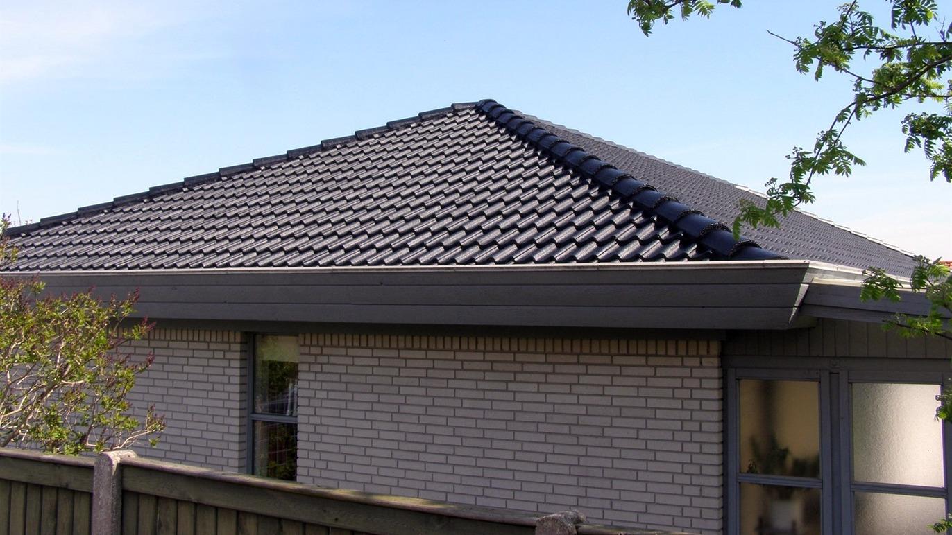 Taget på et hus med mørke tagsten og en klar blå himmel i baggrunden.