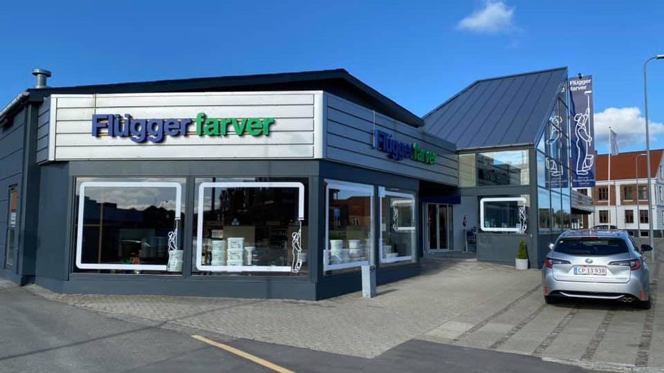 Butik med skiltet "Flügger farver" og store vinduer, beliggende på en gade med en bil parkeret foran.