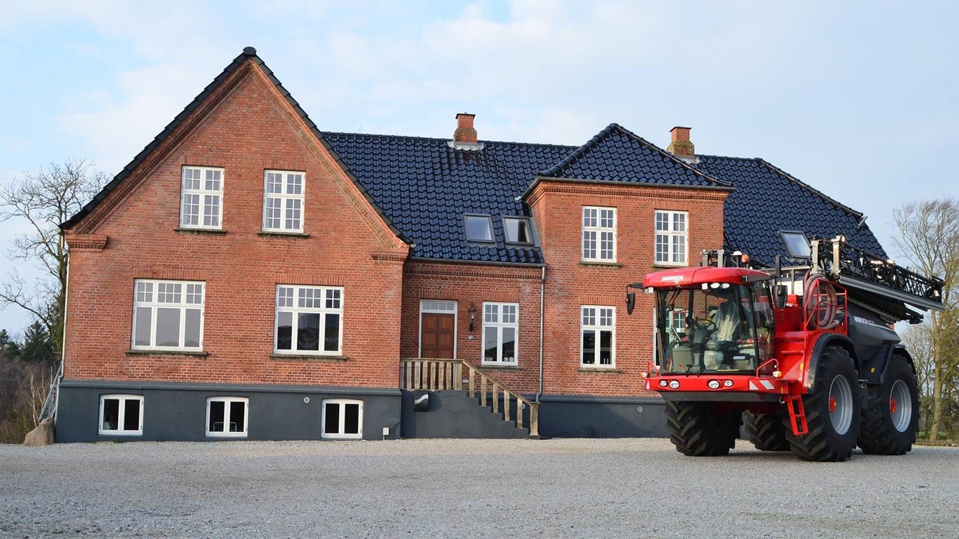 Rødt hus med sort tag og stor sprøjte traktor parkeret foran på grusbelagt område.