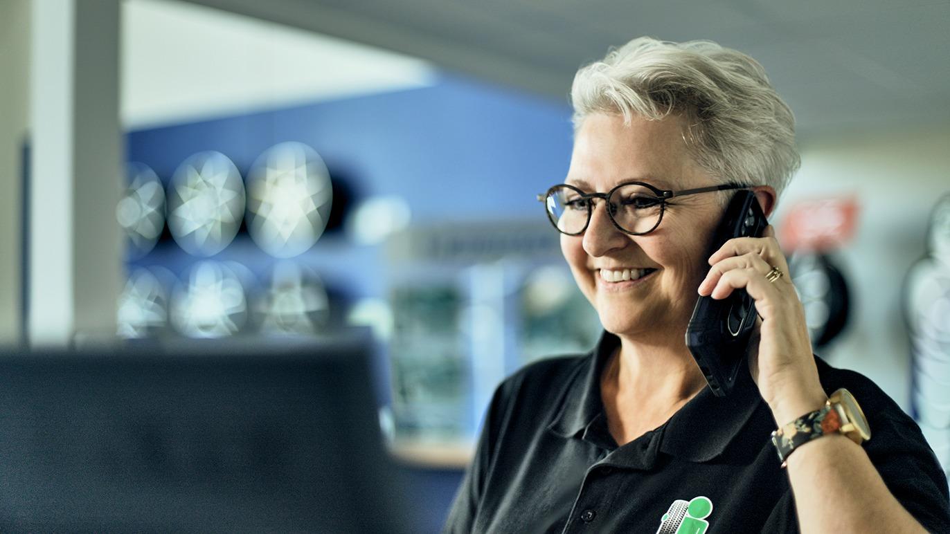 Kvindelig medarbejder taler i telefon og smiler i en moderne kontorindstilling med bilhjul i baggrunden.