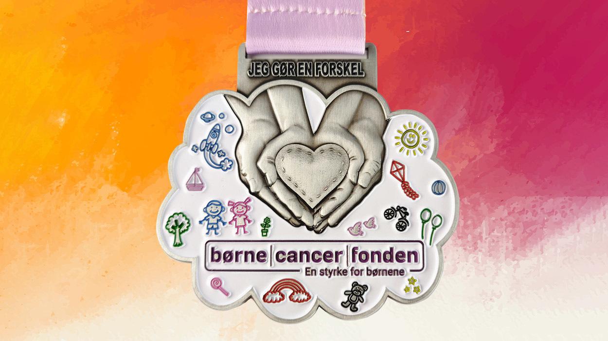 En medalje fra Børnecancerfonden med hænder, der holder et hjerte, og farverige illustrationer af børn og leg.