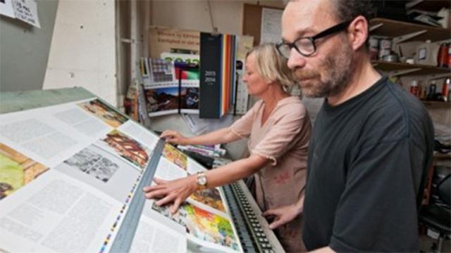 To personer arbejder sammen ved en trykpresse, mens de justerer farverne på tryksager.