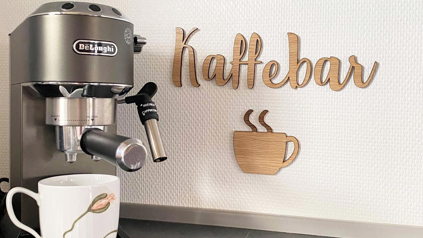 Kaffemaskine ved siden af en træskilt med teksten "Kaffebar" og en kaffekop i forgrunden.
