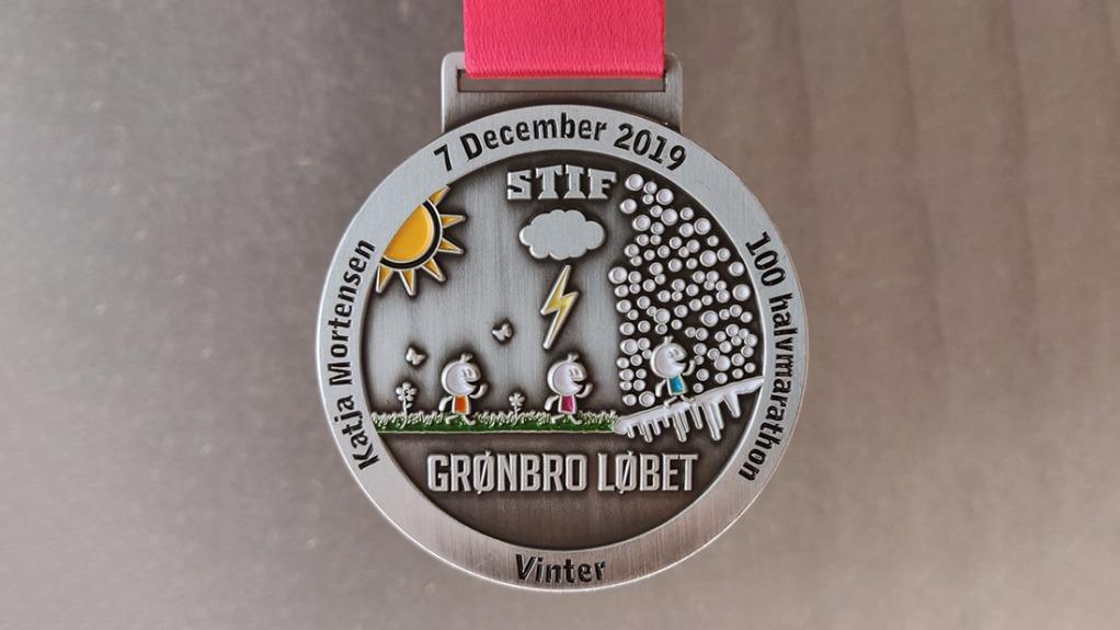 Medalje fra Grønbro Løbet 2019 med illustrationer af løbere, sol, skyer og sne.