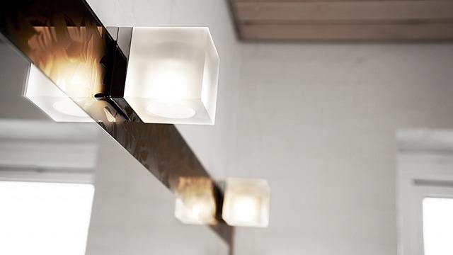 Moderne lampe med firkantede glaskupler hængende fra en metalstang i et lyst rum.