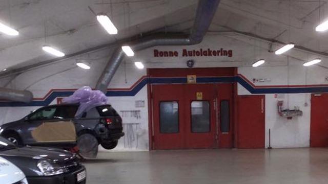 En bil i en autoværksted med en stor lila plastikpose på taget og en rød dør i baggrunden.