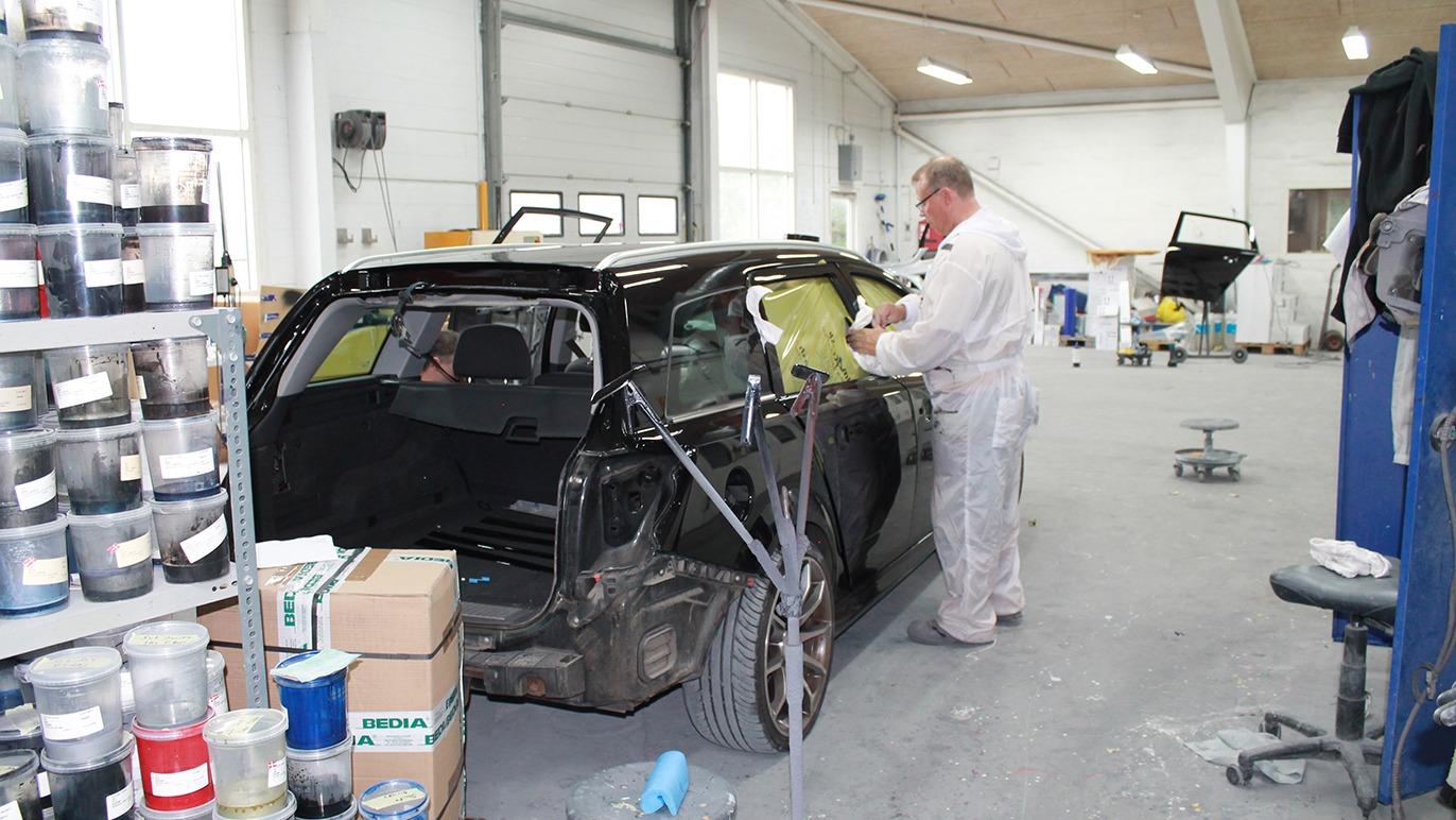 En mekaniker arbejder på en sort bil i et værksted, omgivet af malingsbeholdere og værktøj.
