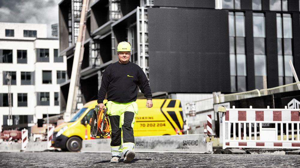En arbejdsmænd i sikkerhedshjelm og arbejdstøj går på en byggeplads med en lastbil i baggrunden.