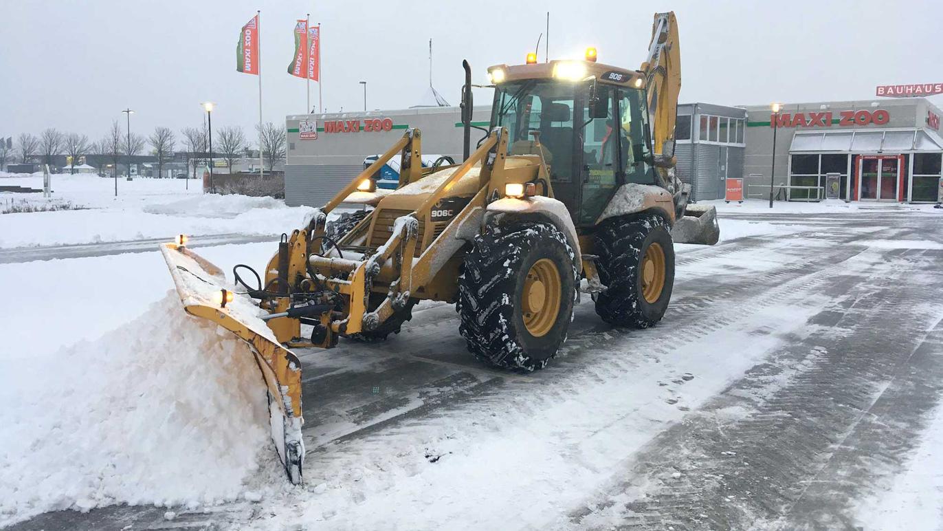En gummihjulstraktor rydder sne fra en parkeringsplads med en sneskraber.