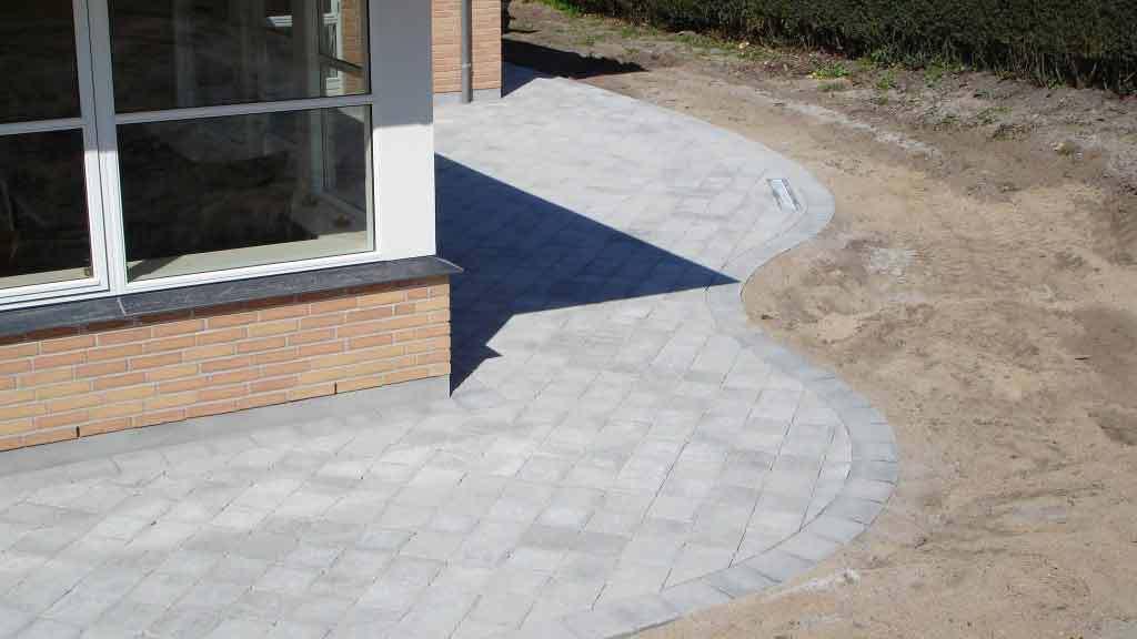 Flisebelægning ved husets facade med en buet kant og sandet område i baggrunden.