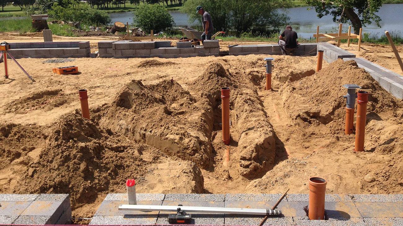 Byggeplads med sand og rør til fundament, arbejdstagere i baggrunden ved en sø.