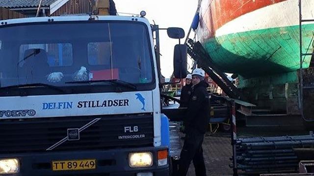 Lastbil ved en båd i en havn, med en person iført sikkerhedsudstyr og båden hævet på en kran.